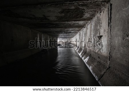 A square concrete drainage tunnel with a light at the end that shines from the turn of the tunnel.
