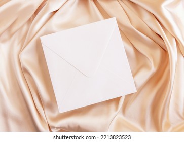 Square Envelope On Beige Silky Fabric Drapery Top View, Mockup. Romantic Scene With Blank White Envelope, Greeting Or Wedding Invitation. 
