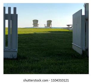 Square format of the backs of twin white Adirondack chairs in sunlight on a green manicured lawn inside an open white gate cast in shade.