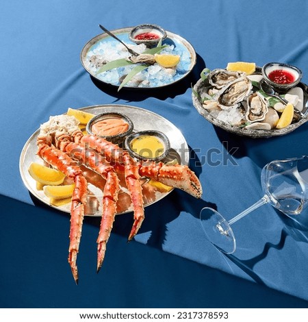 A square frame of a luxurious seafood platter with crab phalanges, lemon slices, sauces, and fresh oysters on ice. Served on a blue tablecloth against a uniform blue backdrop with stark shadows.