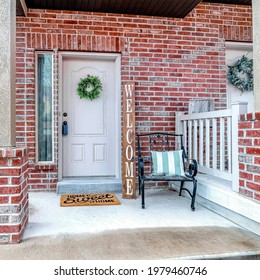 Square Front Of Apartment Building With White Front Door And Red Brick Exterior Wall. White Garage Door And Chair On Snowy Porch Can Also Be Seen At The Facade Of The Residence.