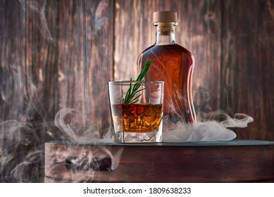 A square glass of whiskey with ice and a sprig of rosemary, and a full bottle of whiskey, wrapped in clouds of smoke, stand on a tray against an old wooden wall. Low key.