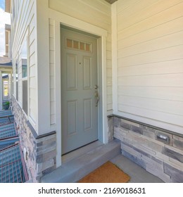 Square Gray Front Door Of A Townhome With Glass Panes And Doormat. Townhome Entrance Exterior With White Vinyl Wood And Stone Veneer Sidings And A View Of Three Basement Window Wells On The Right.