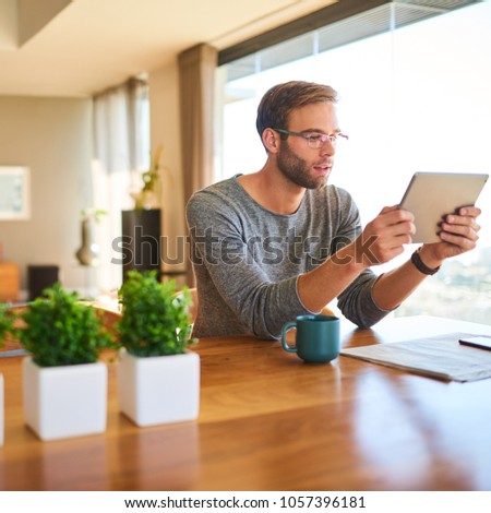 Square image of modern caucasian man sitting at the stunning dining table in his modern home with beautiful large glass windows, while reading his electronic tablet with his morning coffee.