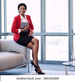 square image of well dressed young black corporate woman wearing a red jacket, holding a digital tablet while sitting on the edge of the couch in a modern business lounge.