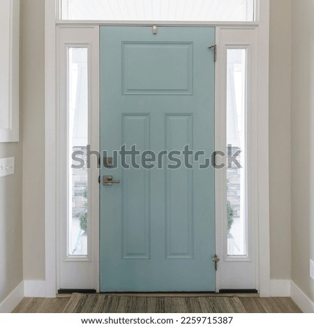 Square Mint front door interior with transom window and sidelights. Interior of a house with light gray walls, wooden flooring and a view of two rooms on both sides with windows.