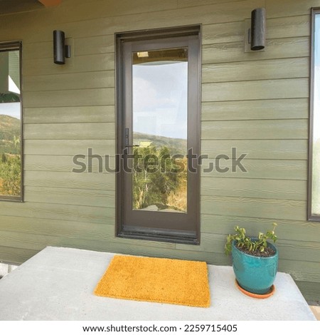 Square Modern traditional exterior of a house with dark green siding. Reflective glass front door of a house and yellow doormat near the potted plant in the middle of two wall lights and windows.
