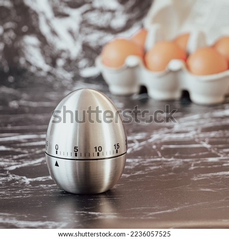 Square photo of silver egg shaped egg timer in the foreground with open box of eggs in the background, against black and white marble effect backdrop.