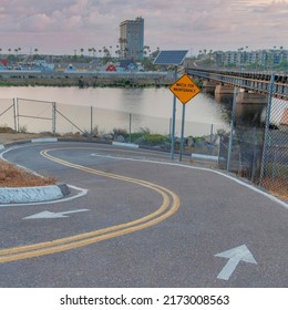 Square Puffy Clouds At Sunset Two-way Curved Downhill Bike Path At Oceanside, California. Bike Trail Near The River With Watch For Maintenance Sign And Solar Panel On A Post And A City View