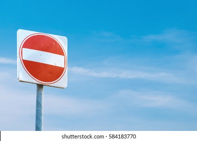 A square red sign with a white bar indicating 'NO ENTRY' on a grey metal post against a blue cloudy sky.