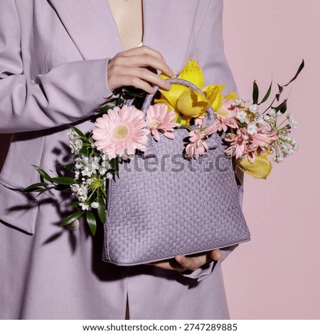 Square studio shot of a trendy spring fashion concept. An unrecognizable woman wearing a stylish pastel violet suit holds a matching woven leather handbag overflowing with vibrant yellow tulips, pink gerberas, and delicate white flowers. Set against a soft pink background with hard shadows, this elegant aesthetic is perfect for spring lookbooks, Mother's Day campaigns, and creative floral fashion concepts