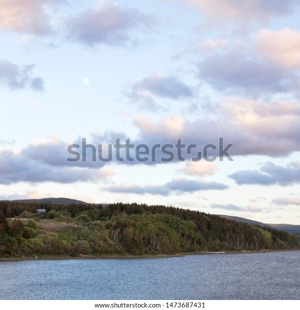 Square view of a\
pure three-quarter moon above a landscape of greens and browns in\
front of a lake inlet with a barn and houses in the distance, near\
Cheticamp, Nova Scotia