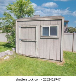 Square White puffy clouds Backyard shed with window near the large rocks and trees on a green lawn. Shed exterior with light gray wood claddings against the vinyl fence and clear blue sky.
