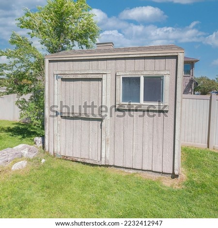 Square White puffy clouds Backyard shed with window near the large rocks and trees on a green lawn. Shed exterior with light gray wood claddings against the vinyl fence and clear blue sky.