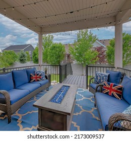 Square White Puffy Clouds Cozy Outdoor Deck With Fire Pit Table And Woven Sofas With Blue Cushions. There Is A Blue Carpet Under The Furnitures And An Outdoor String Lights On The Ceiling