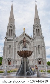 St Anne De Beaupre, Quebec, Canada - June 24, 2010: The Front Of The Basilica Of Sainte-Anne-de-Beaupré Church With Statue Of Saint Anne By Canadian Sculptor Émile Brunet On A Fountain.