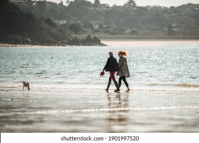 St Brelades Bay Channel Island April 2020 Jersey Two Ladies Walking Dog Sky Beautiful Seashore Sand Beach In Morning Reflection On Wet Sand Hotel Clouds Horizon Reflecting Sun On Ocean Uk Holiday