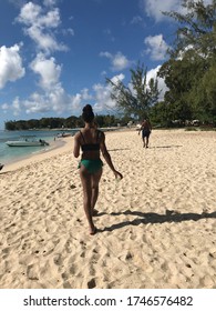 St. James, Barbados - April 17, 2019: A Rear View Of A Black Woman In A Bikini Walking On The Beach In The Foreground With A Black Man In A Bathing Suit In The Background 