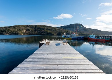 St. John's, Newfoundland, Canada-February 2022: A Long Grey Wooden Wharf With Multiple Chairs Positioned At The End Of The Pier Overlooking A Clear Blue Ocean. Colorful Boats Are Moored On A Dock.