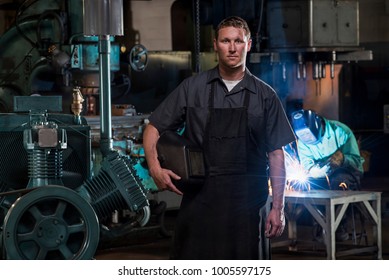St Louis, Missouri, United States-circa 2015-Portrait of male welder with mask standing near compressor generator in dirty manufacturing factory plant with man welding in background, sparks flying.