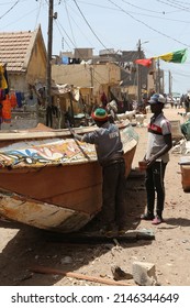 ST LOUIS, SENEGAL – MARCH 19, 2022: Street In Saint Louis City, Senegal, Africa. Boat And People In Traditional National Costumes, Dresses. Saint Louis Landmark, Monument. Senegalese Fishing Ship