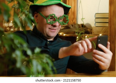 St Patrick Day Concept. Man Celebrating Saint Patricks Day At Home. Man In Hat And Green Glasses Communicating With Friends Using Smartphone.