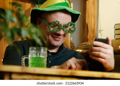 St Patrick Day Concept. Man Celebrating Saint Patricks Day At Home, Having Video Call. Man In Hat And Green Glasses Communicating With Friends Using Smartphone.