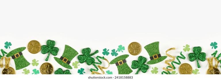 St Patricks Day bottom border of green shamrocks, leprechaun hats, gold coins and ribbon. Top down view over a white banner background with copy space.