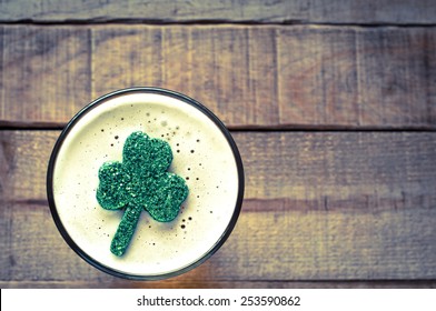 St. Patricks Day, Irish Shamrock Floating In Frothy Beer Mug On Rustic Wood Board Background With Room Or Space For Copy, Text, Your Words.  Horizontal Faded Filter Instagram, Tint, Dark And Moody 