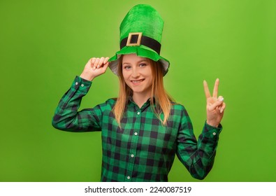 St Patricks Day leprechaun girl in green festive irish hat and checkered plaid shirt showing peace gesture v sign with two fingers and looking at camera isolated on green background.