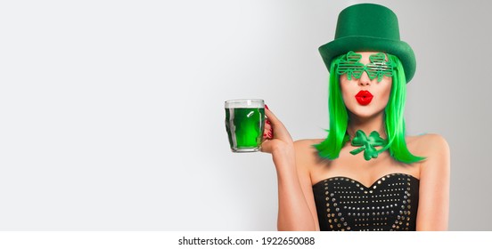 St. Patrick's Day leprechaun model girl in green hat and costume, holding mug of Green Beer pint over gray background, surprised emotion. Patrick Day pub party, celebrating. Glass of Green beer. 