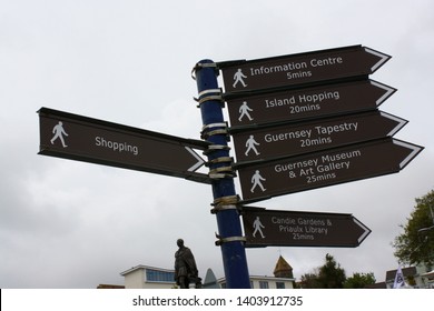 St. Peter Port, Guernsey, Channel Islands - May 2, 2019: Sign Providing Information To Various Location And The Estimated Walking Time In Minutes With The Monument To Prince Albert In The Background