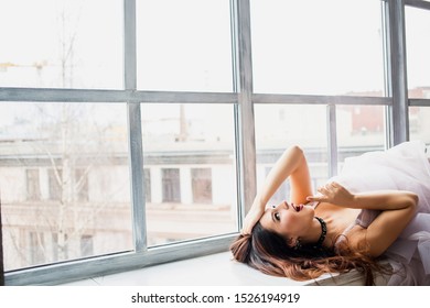 
St. Petersburg, Russia - September 25, 2019: A Beautiful Young Girl In A Lilac Dress, The Bride In An Unusual Way, Temporarily Lips Sits / Lies On A Window Sill Near A Large Window. Rock And Roll And