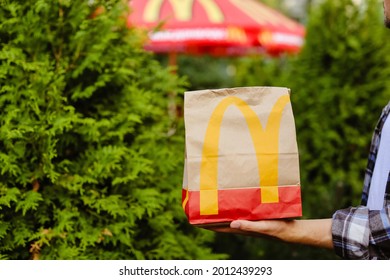 St Petersburg,Russia 18.07.2021 McDonald's Fast Food Restaurant With Delicious Unhealthy.Courier Man Holds Paper Bag With Parcel Green Background Of Trees In Garden Summer.Delivery Of Order In Package