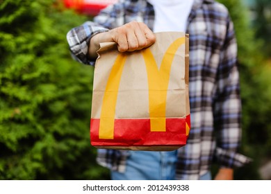 St Petersburg,Russia 18.07.2021 McDonald's Fast Food Restaurant With Delicious Unhealthy.Courier Man Holds Paper Bag With Parcel Green Background Of Trees In Garden Summer.Delivery Of Order In Package