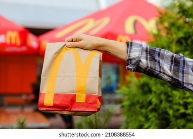 St Petersburg,Russia 18.07.2021 McDonald's Fast Food Restaurant With Delicious Unhealthy.Courier Man Holds Paper Bag With Parcel Green Background Of Trees In Garden Summer.Delivery Of Order In Package