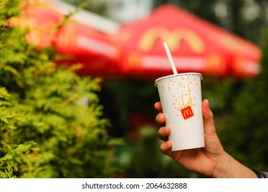 St Petersburg,Russia 18.07.2021 White Cardboard Glass With Plastic Tube Lemonade Juice Yellow Red McDonald's Logo.Fast Food Restaurant McDonald Street.Mug Drink In Hand Man Background Green Plant