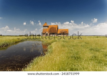 St Thomas Becket Church in Fairfield Romney Marsh Kent. A lone ancient chapel sits in grass and marshland with sheep grazing around and watercourses and dykes beside.