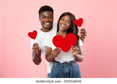 St. Valentines Day Concept. Portrait Of African American Couple In Love Holding Red Paper Heart Cards, Smiling Guy Standing Behind Woman's Back, Looking At Camera, Isolated On Pink Studio Background