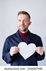 St. Valentine's Day Symbols: Portrait Of Young Bearded Male Construction Worker Standing By The Wall Holding White Paper Heart Shape. Romance Or Relationships Concept. High Quality Image