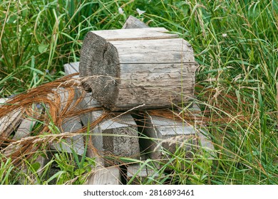 A stack of aged, textured wooden logs with coils of rusted metal wire resting in a lush green field of tall grass and wildflowers.