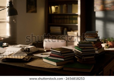 Stack of assorted books arranged on desk in home office with sunlight streaming through window, bookshelf filled with volumes in background, workspace suggesting academic or literary setting