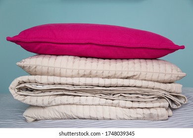 A Stack Of Beige Folded Blankets And A Red Pillow Lie On The Net-gray Bedspread In The Room. Gray Blue Wall In The Background