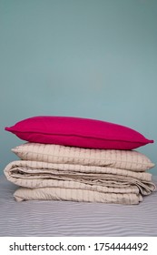 A Stack Of Beige Folded Blankets And A Red Pillow Lie On The Net-gray Bedspread In The Room. Gray Blue Wall In The Background