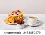 Stack of Belgian waffles, berries and cup of tea: delicious summer breakfast on marble table. Homemade waffle and mix of raspberries and blueberries as topping. Sweet dessert on white dishes