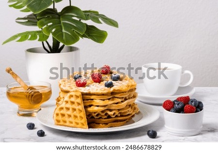 Stack of Belgian waffles, berries, honey and cup of tea: delicious summer breakfast on marble table. Homemade waffle and mix of raspberries and blueberries as topping