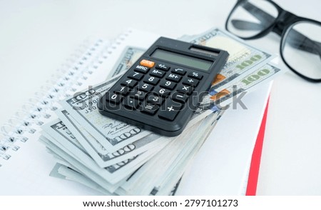 Stack of bills rests beside a calculator. Black glasses sit nearby on a clean white desk. Focused financial calculations or budgeting is implied. Ideal for business, finance