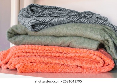 A stack of blankets folded on a modern white shelf, including a bright orange plush throw and gray blankets. The combination of colors and textures creates a cozy home vibe with a balance of warmth