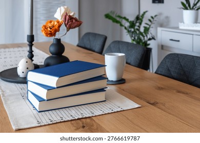 Stack of blue books and a cup on a wooden table in a home interior. Decorative elements like a candle holder and vase are visible in the background. Suitable for themes like reading, studying, learning and everyday life at home.