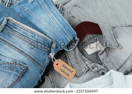 Stack of blue jeans, shirts and tag with inscription second hand. Second hand clothing shop. Circular fashion, eco friendly sustainable shopping, thrift stores concept. Top view over woman outfit